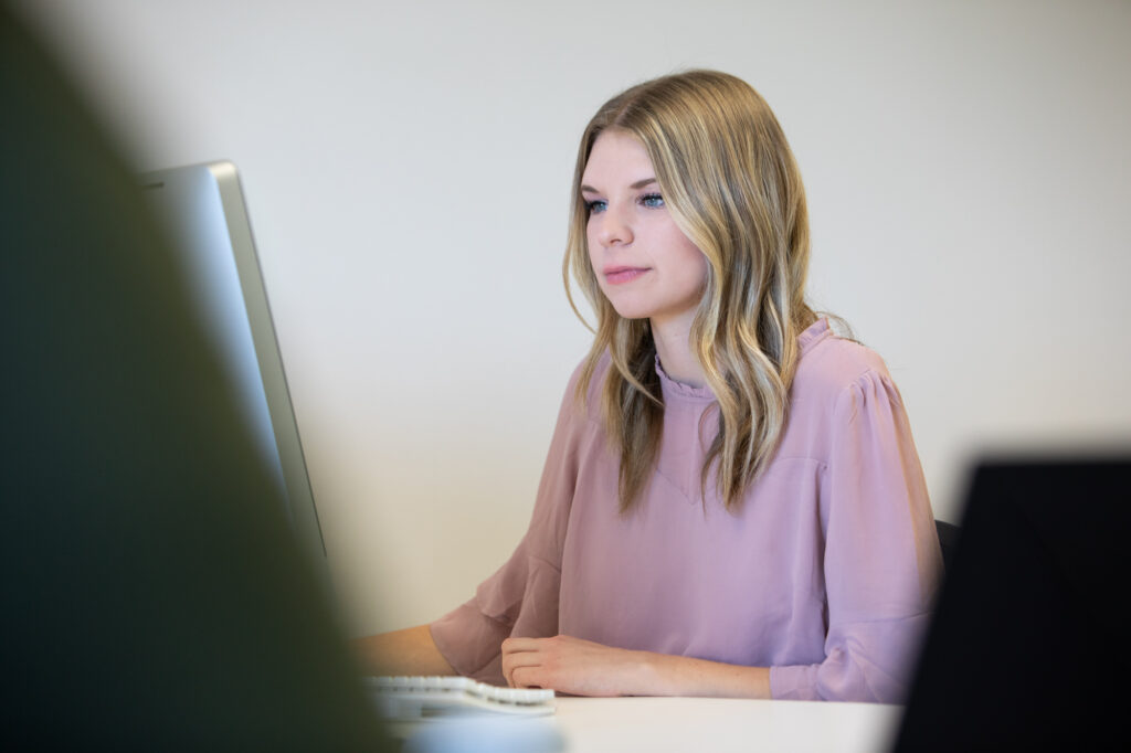 A focused professional woman working at a desktop computer in a modern office. She is wearing a light pink blouse, with a calm and thoughtful expression, exemplifying a productive work environment.