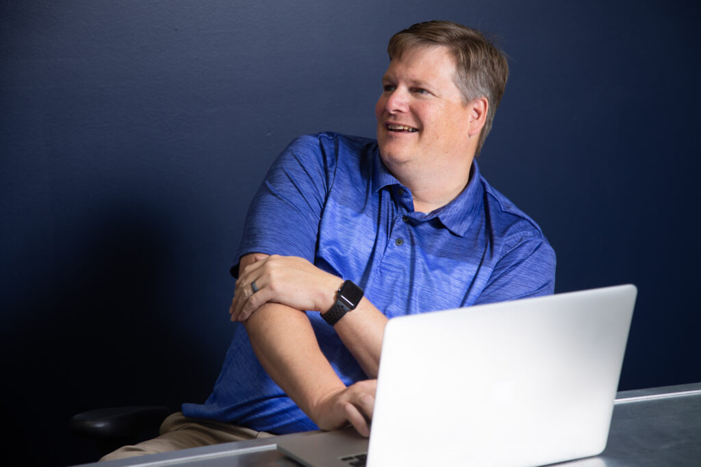 A smiling professional man in a blue polo shirt sitting at a desk with a laptop, engaging in a conversation. The navy blue wall background adds a modern and professional touch.