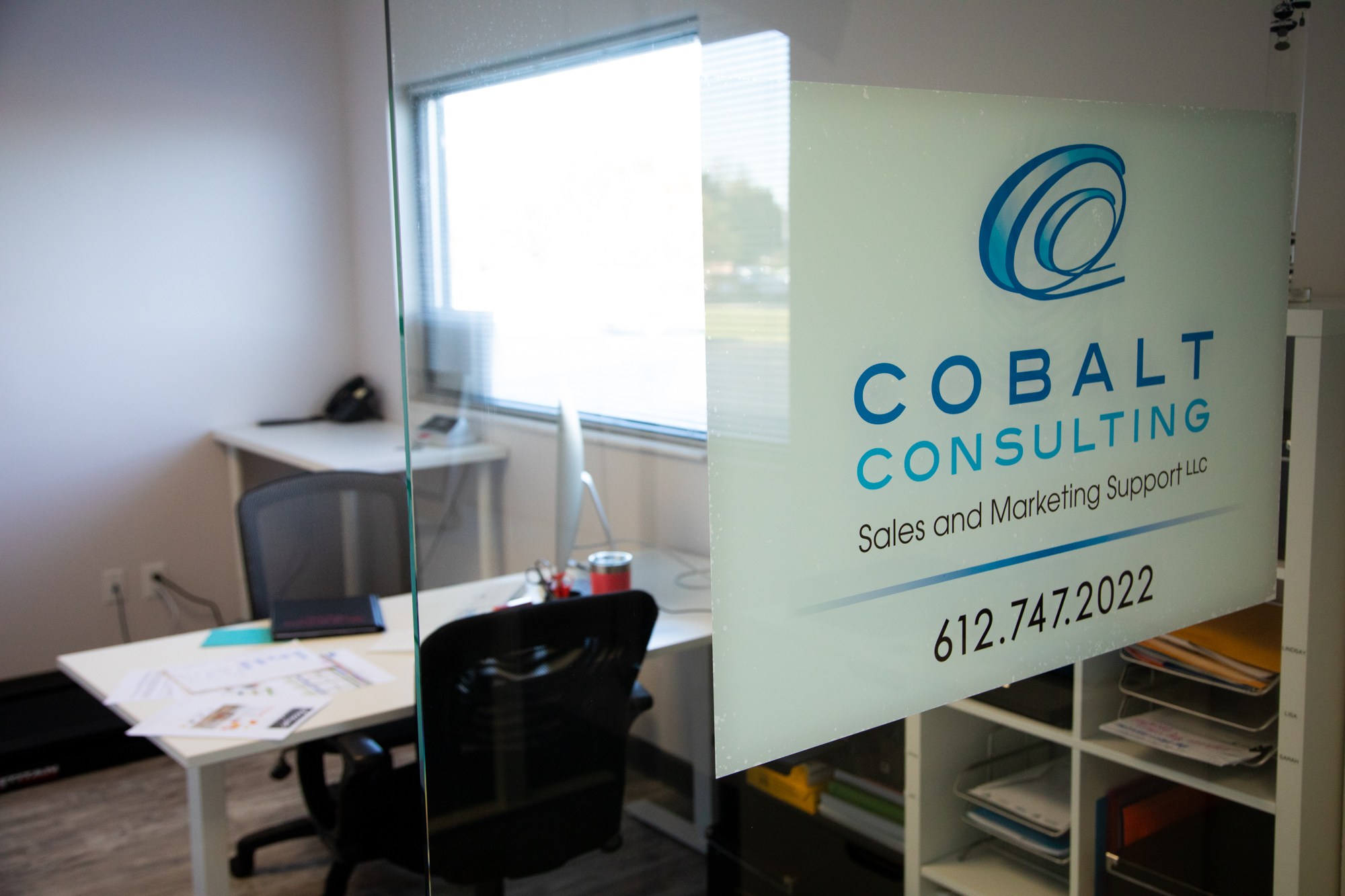 Office view with a glass door featuring the Cobalt Consulting logo, tagline "Sales and Marketing Support LLC," and contact number "612.747.2022." Inside, a tidy workspace with a desk, chair, and documents emphasizes a professional environment.