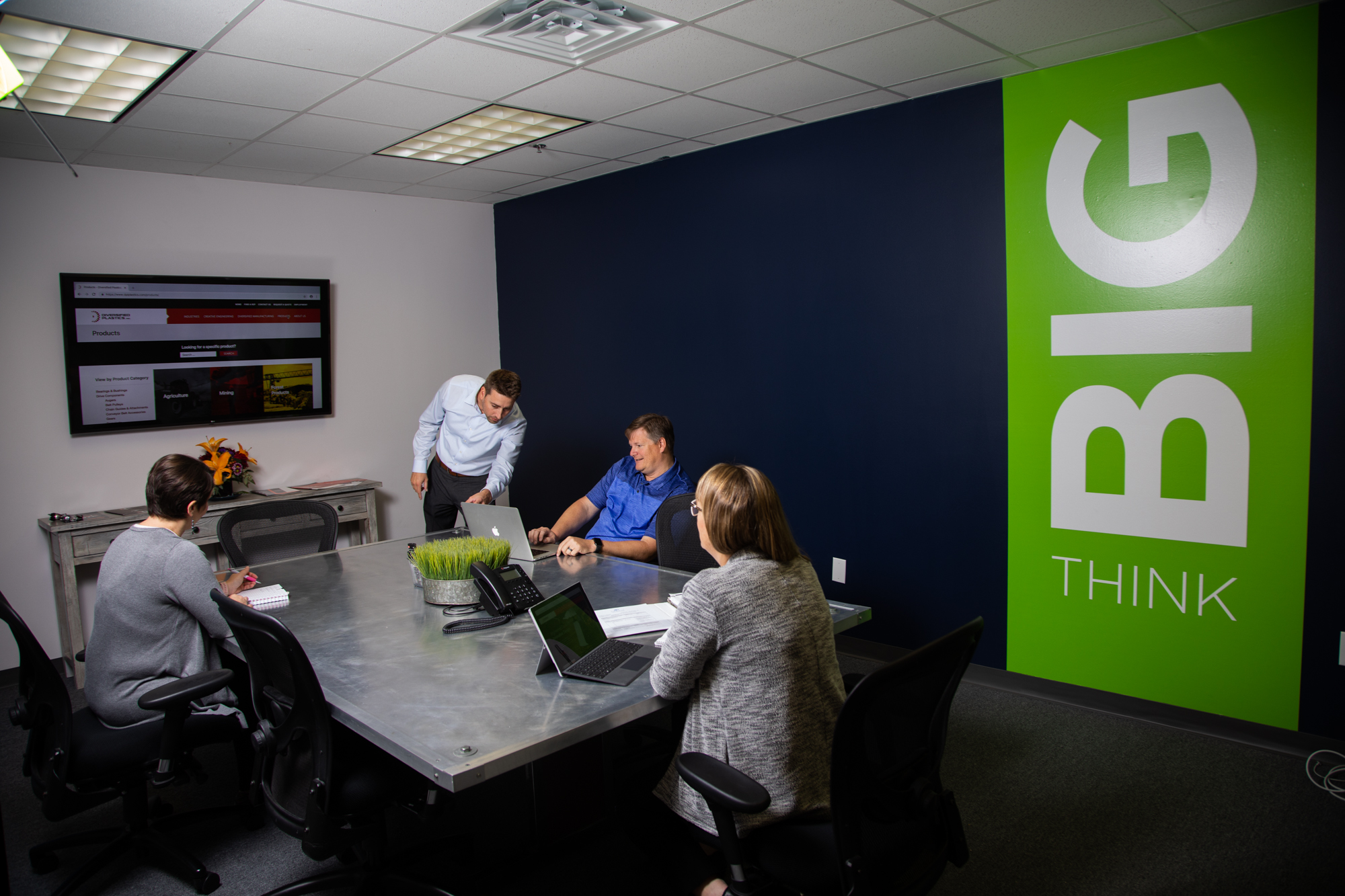 A professional office setting with four individuals collaborating around a modern conference table. A man is presenting on a laptop, while others are engaged in discussion. A wall features the phrase "THINK BIG" in bold green and white letters, emphasizing creativity and innovation.