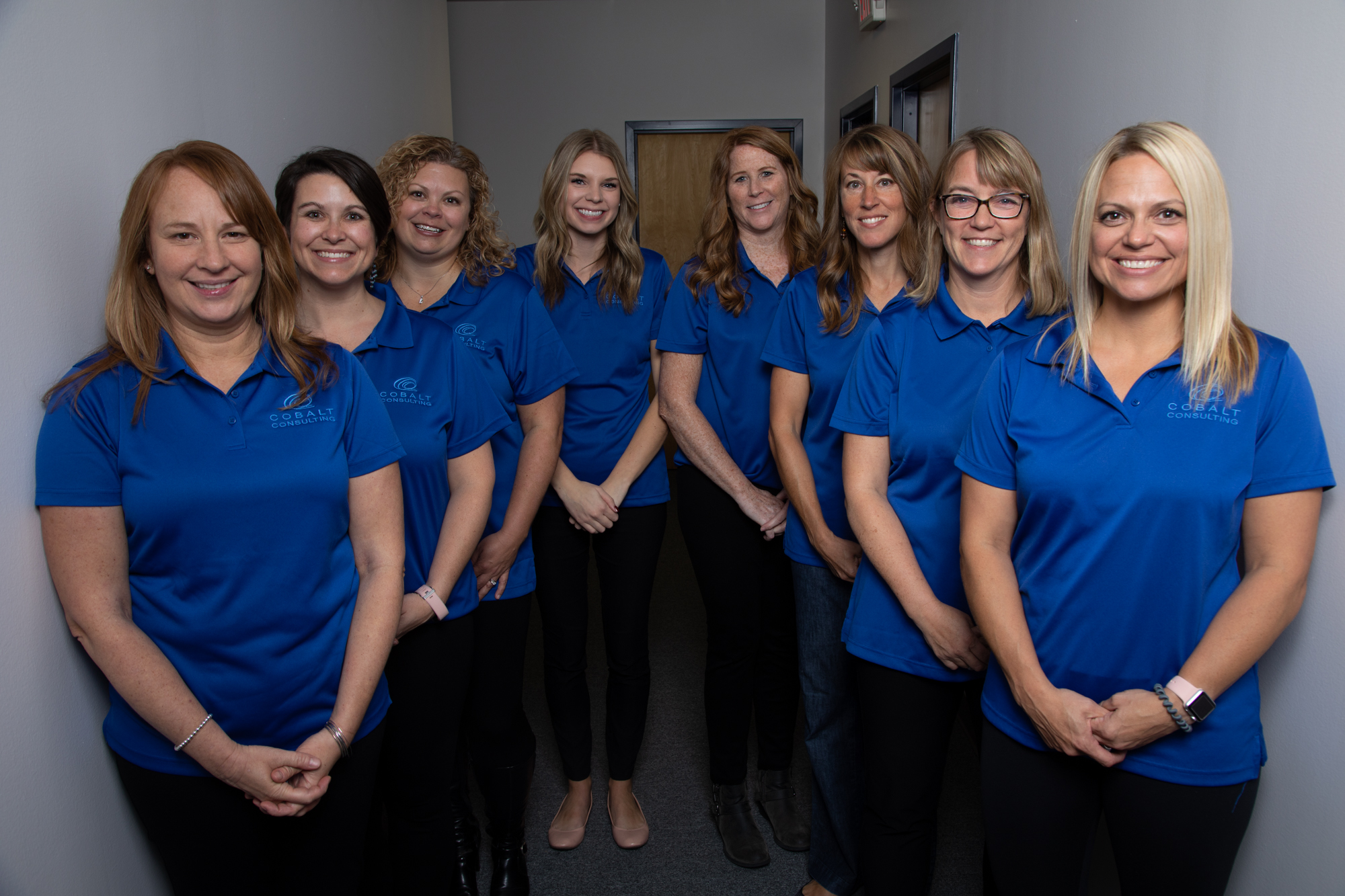 A team of eight professionals from Cobalt Consulting standing together in a hallway. They are all wearing matching blue polo shirts with the company logo, showcasing unity and professionalism.